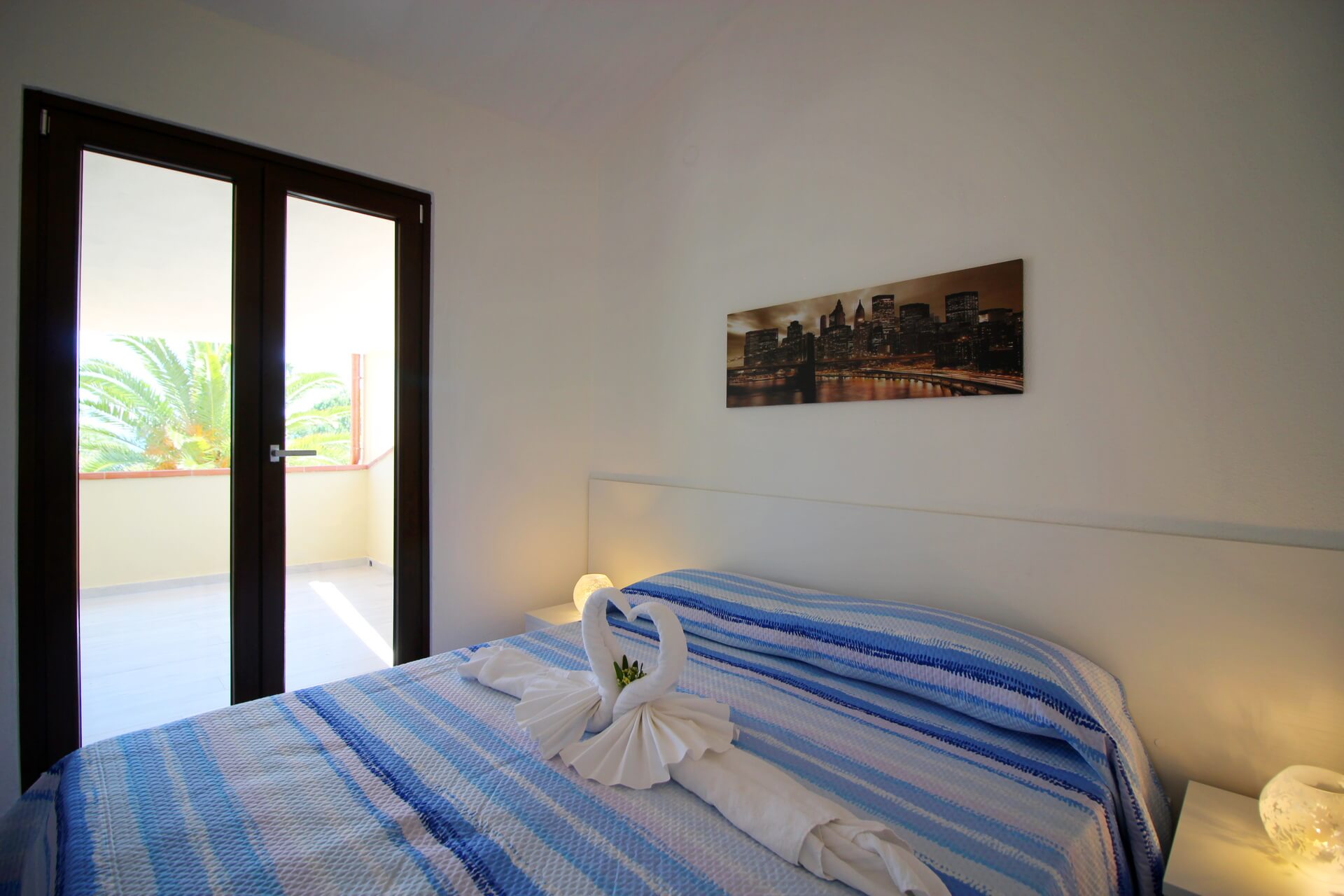 Letto matrimoniale con lenzuola a righe blu e bianche, porta-finestra che si apre sulla veranda